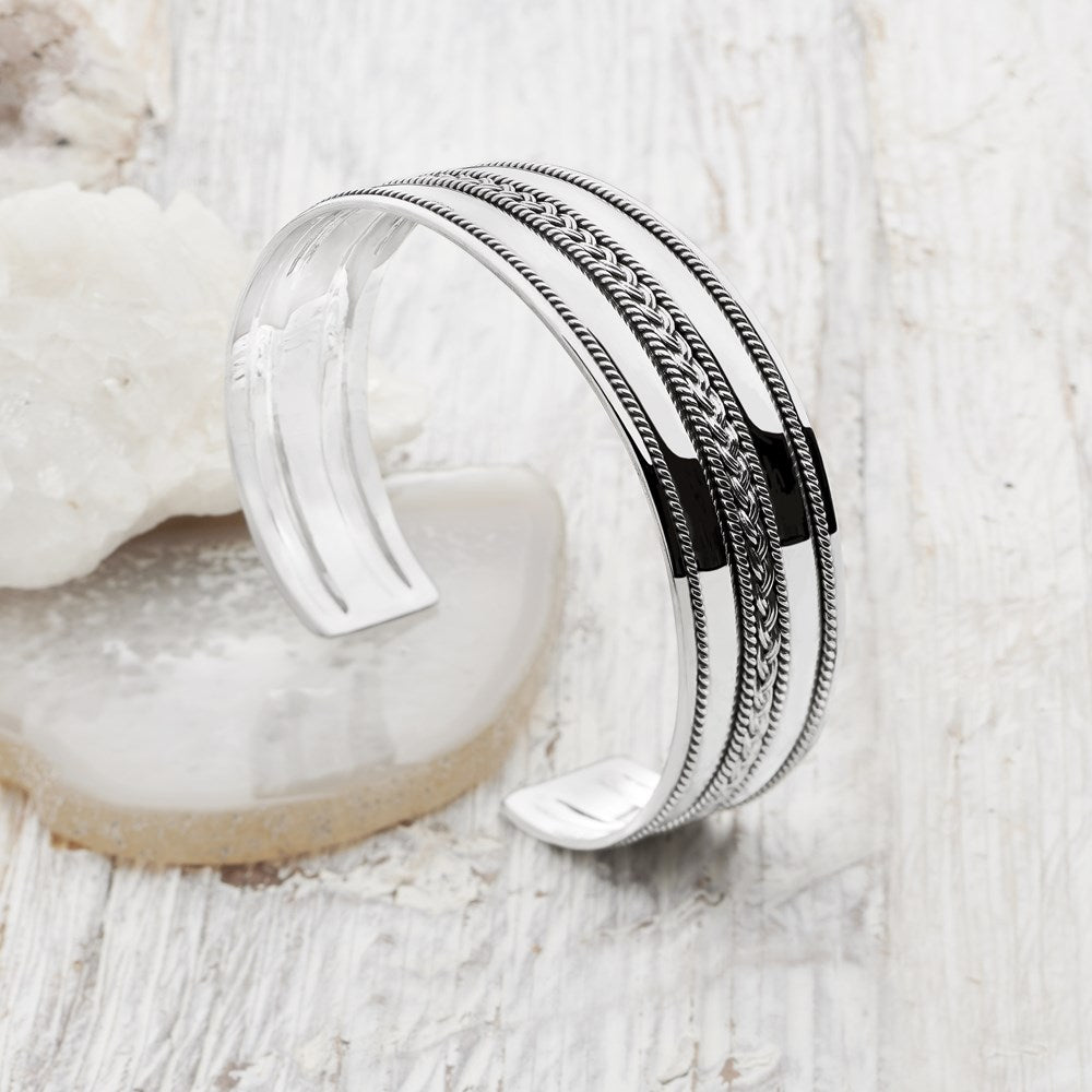 A silver bangle bracelet with a woven pattern and oxidised finish displayed on a white surface.