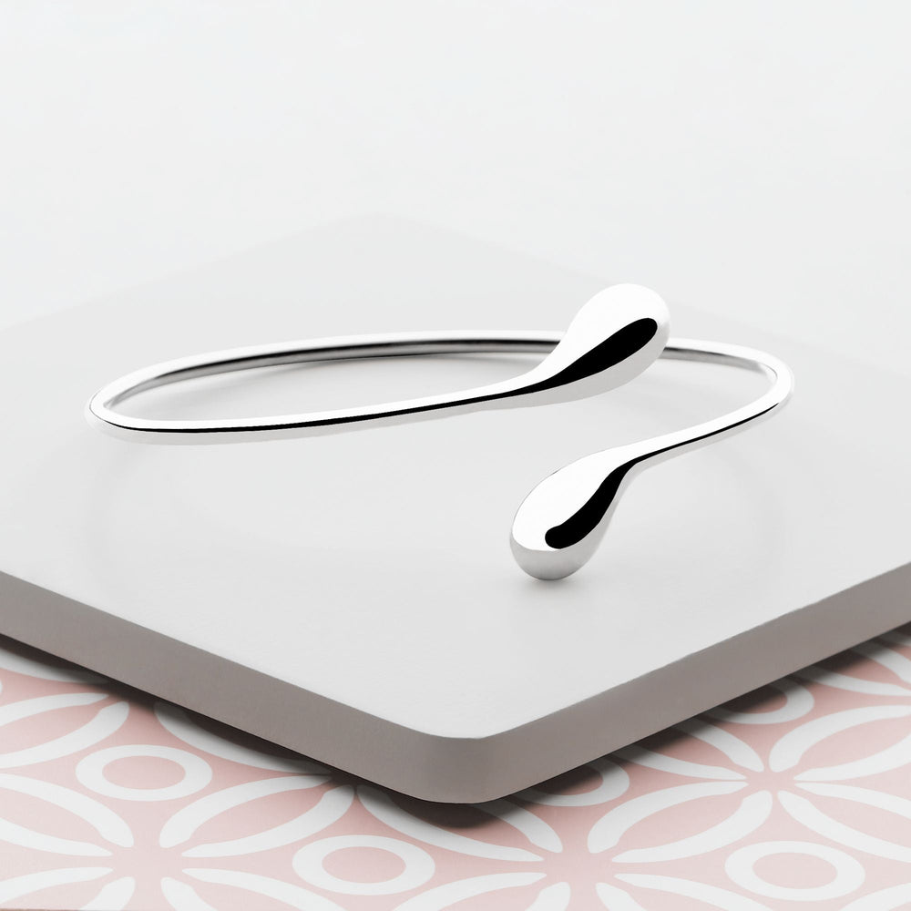 A silver bangle bracelet with a design that ends in two raindrop shapes, on neutral background. 