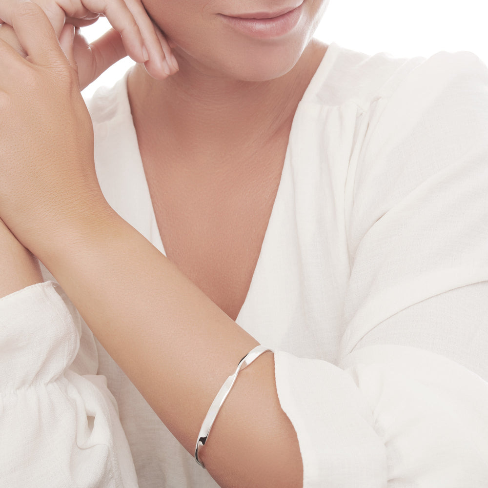 A silver bangle bracelet with a high polish finish, displaying a smooth, ribbon-like design on model.