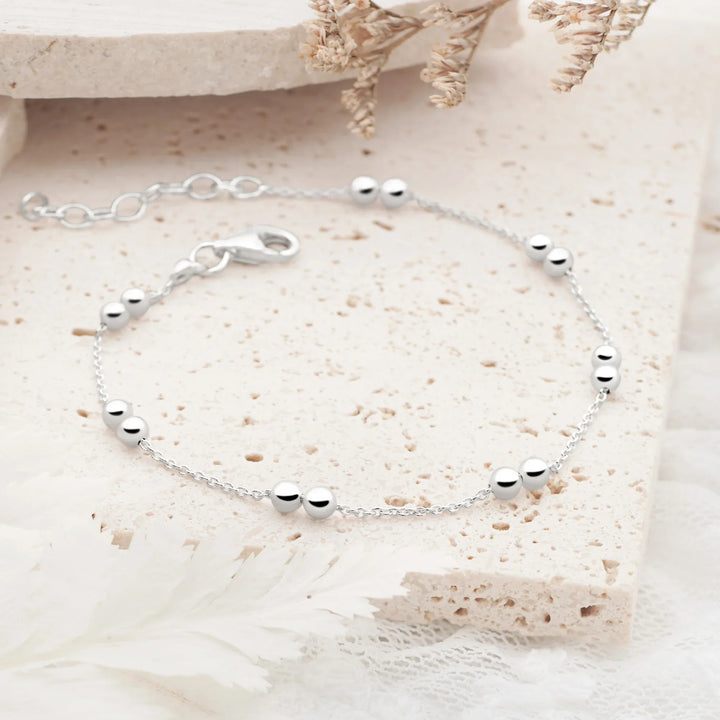 A silver bracelet with twin beads on a classic link chain, displayed on a white surface with a patterned background.