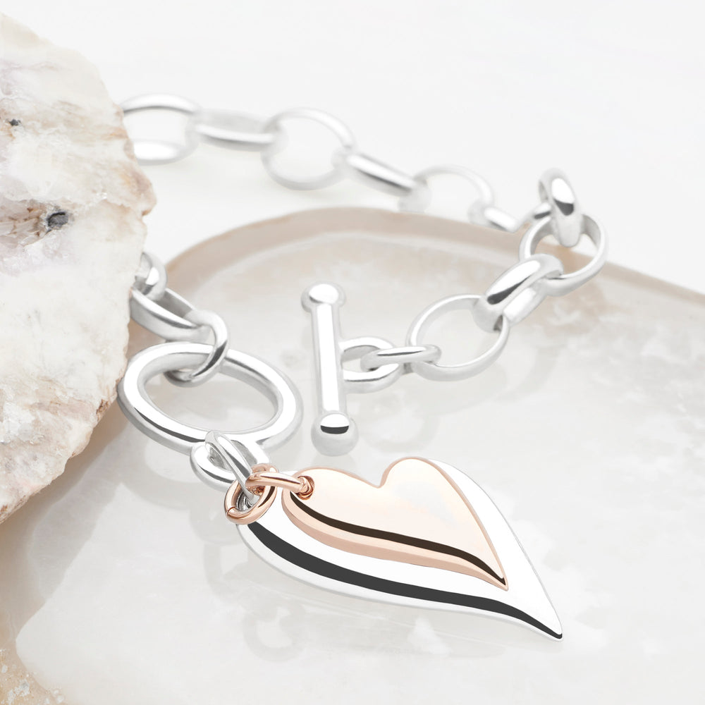 A silver bracelet with a rose gold-plated heart charm, featuring a T-bar fastening on a chain.