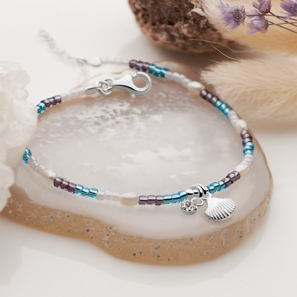 A bracelet with creamy white pearls, colorful beads, and a silver shell charm, displayed on a beach-themed setting.