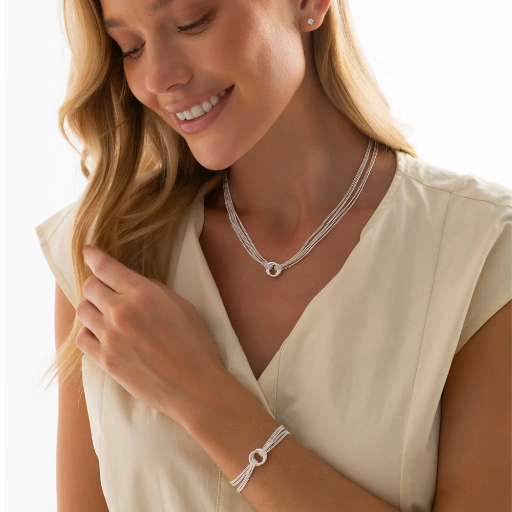 A woman wearing a silver necklace and bracelet from the Links of Love Silver Set collection. The necklace is long and has multiple layers of loops and chains, while the bracelet is also with loops and chains.