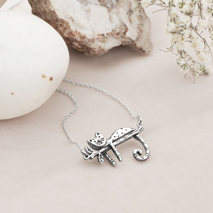 A silver necklace with a pendant in the shape of a relaxed kitty-cat, lying on a branch.