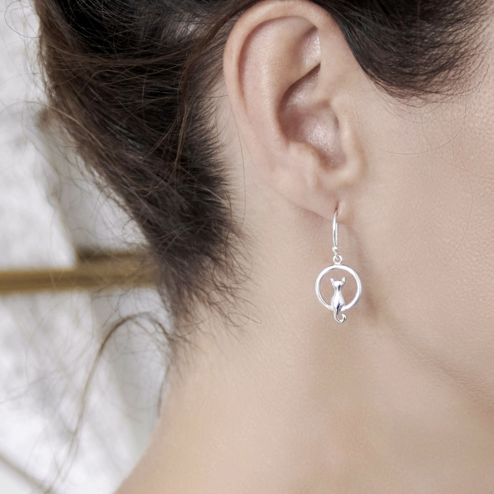 A pair of sterling silver earrings in the design of a cat sitting inside a circle with a tail loop, high polish finish on model.
