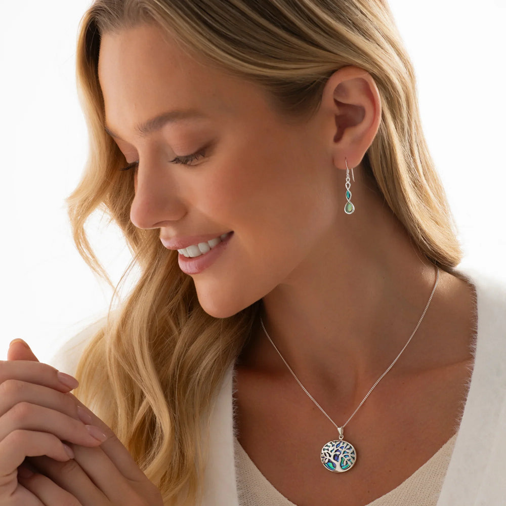 A silver pendant with a Tree of Life design and an abalone shell pattern in the center, held by its loop on a person's neck.