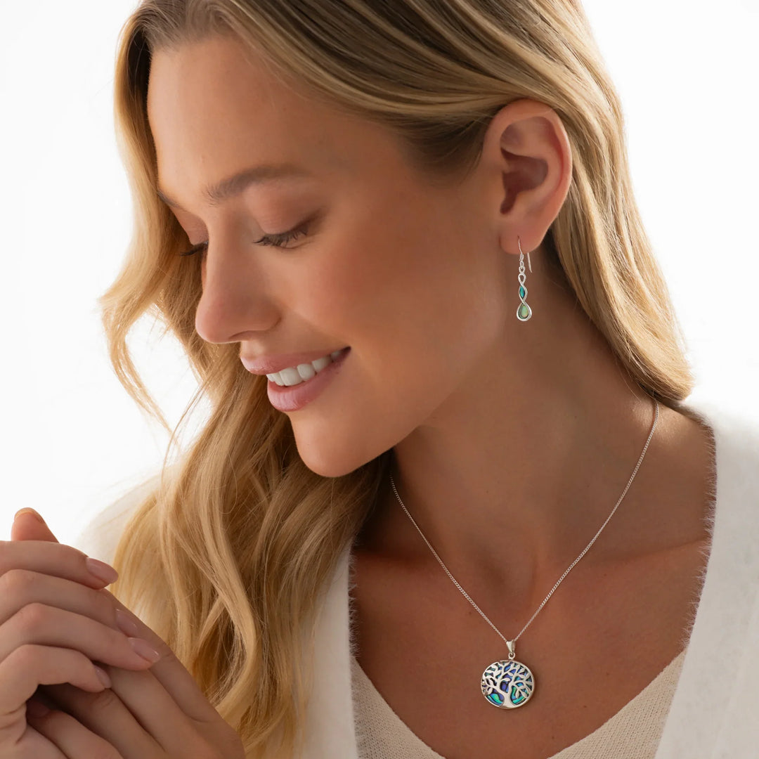 A silver pendant with a Tree of Life design and an abalone shell pattern in the center, held by its loop on a person's neck.