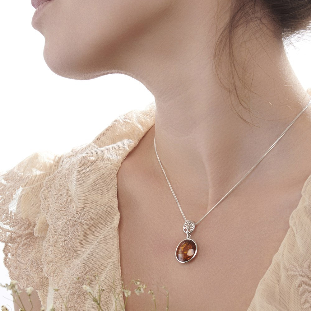 A female model wearing a silver pendant with a tree of life design and a cabochon of amber swinging below it, attached to a silver chain.