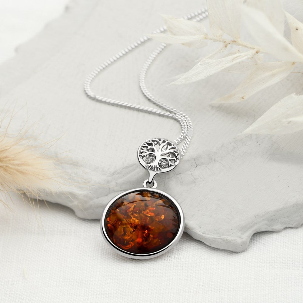 A silver pendant with a tree of life design and a cabochon of amber swinging below it, attached to a silver chain.