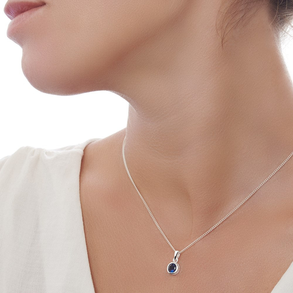 A model wearing a sterling silver pendant with a blue sapphire cubic zirconia set in a round, silver frame, attached to a 40cm curb chain.