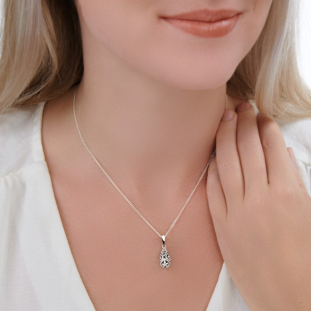 A silver pendant with a teardrop shape and Balinese inspired design, attached to a silver curb chain on a woman model. 