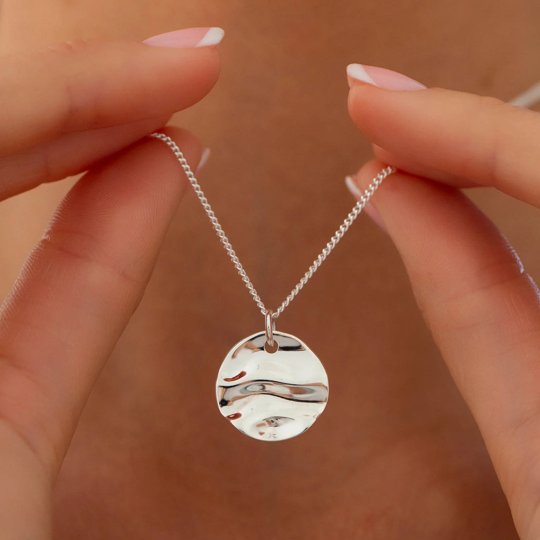 Silver necklace with a round pendant held by a hand against a beige background